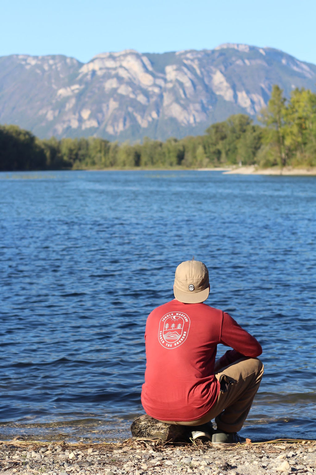 A man wearing a red long-sleeve shirt with a circular white graphic on the back, a beige cap, and khaki pants sits on a rock at the edge of a blue lake with mountains in the background.